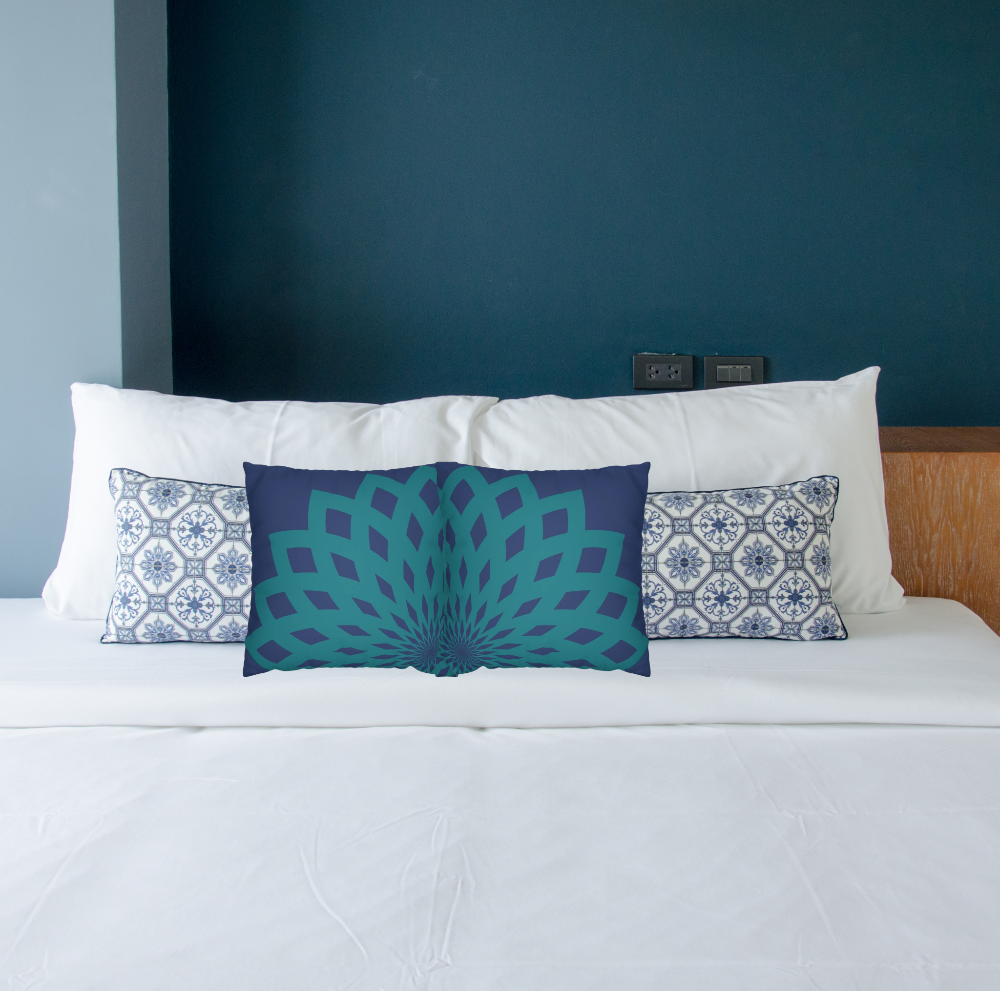 Blue and teal decorative pillow shown on a bed with contrasting printed pillows