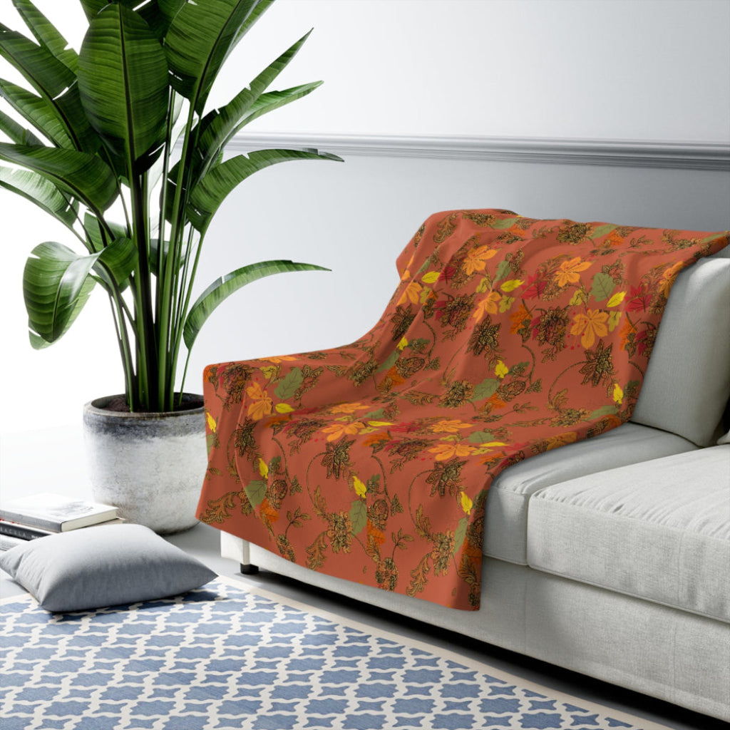 Floral-patterned orange blanket with green and yellow leaves draped over a white sofa in a living room setting with a plant and rug.