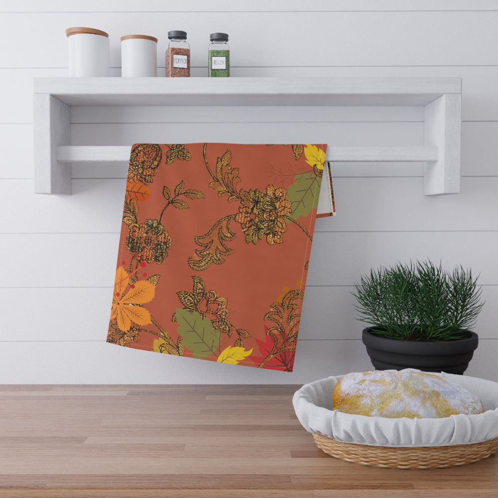 Deep orange background with bronze floral pattern and multi colored fall leaves on a kitchen tea towel, shown hanging in a kitchen