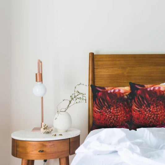 A styled bedroom scene featuring two red and orange textured floral throw pillows on a bed with white sheets, next to a wooden nightstand with a copper and white table lamp.