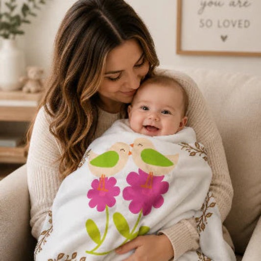 A smiling baby wrapped in a white blanket with a colorful floral and bird print, held by a woman on a beige couch in a cozy living room setting.