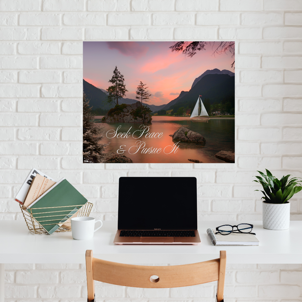 Desk with a laptop, mug, and books, featuring a scenic wall art print with a sailboat and mountain landscape with the text Seek Peace and Pursue it.