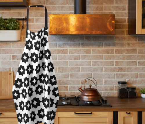A black and white farmhouse style apron with geometric print shown hanging next to an elegant copper range hood/vent 