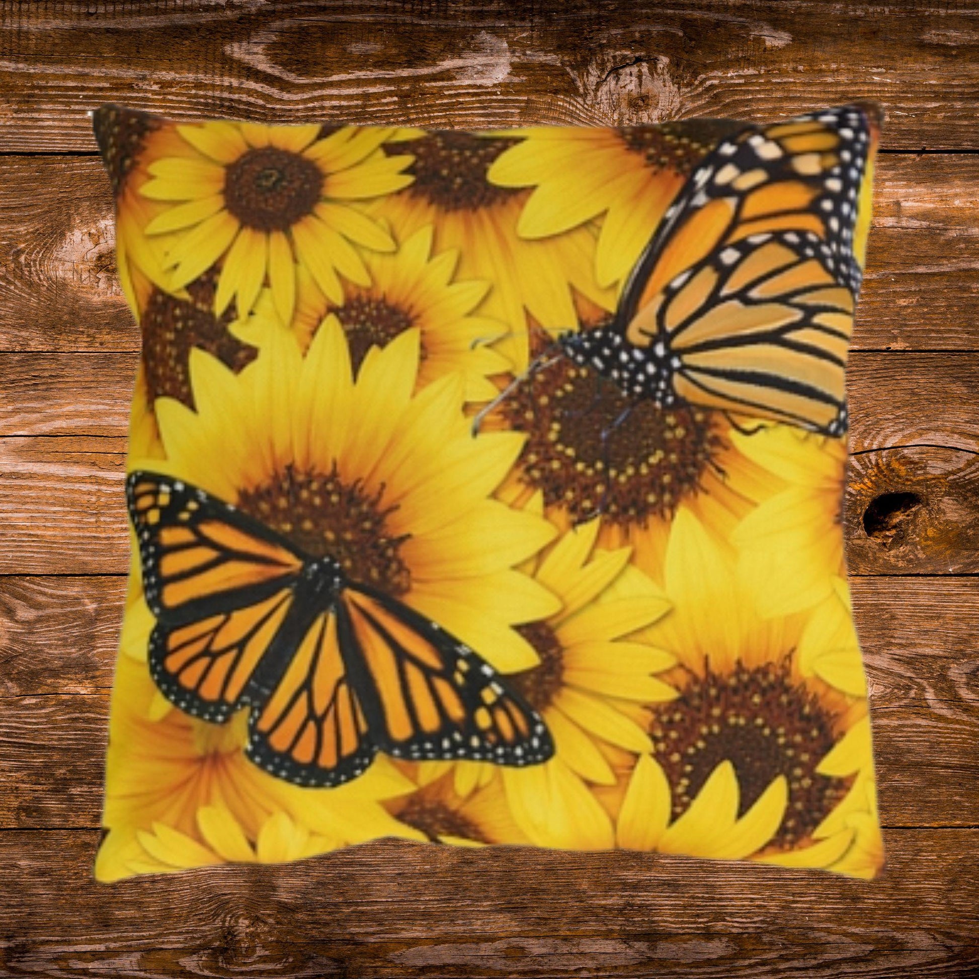 A square throw pillow featuring a vibrant print of yellow sunflowers and orange-and-black monarch butterflies, displayed flat on a rustic brown wooden plank background.