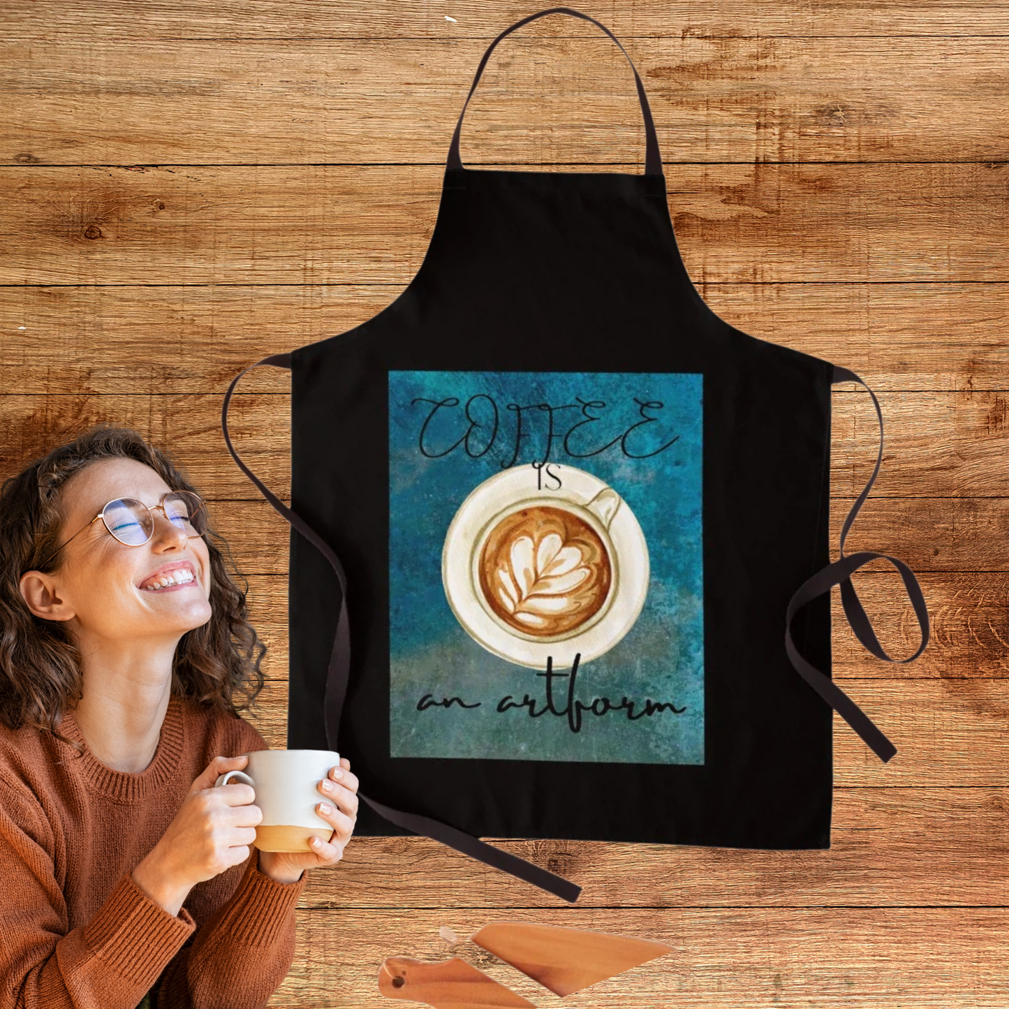 picture shows a black apron with teal graphic background showing a cup of cappuccino  with a woman in front of it sipping coffee