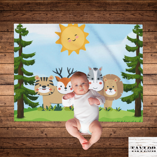 A baby wearing a white bodysuit laying on a colorful printed blanket featuring playful animals, trees, a sun, and a blue sky. The blanket is laid flat on a rustic brown wood plank floor.