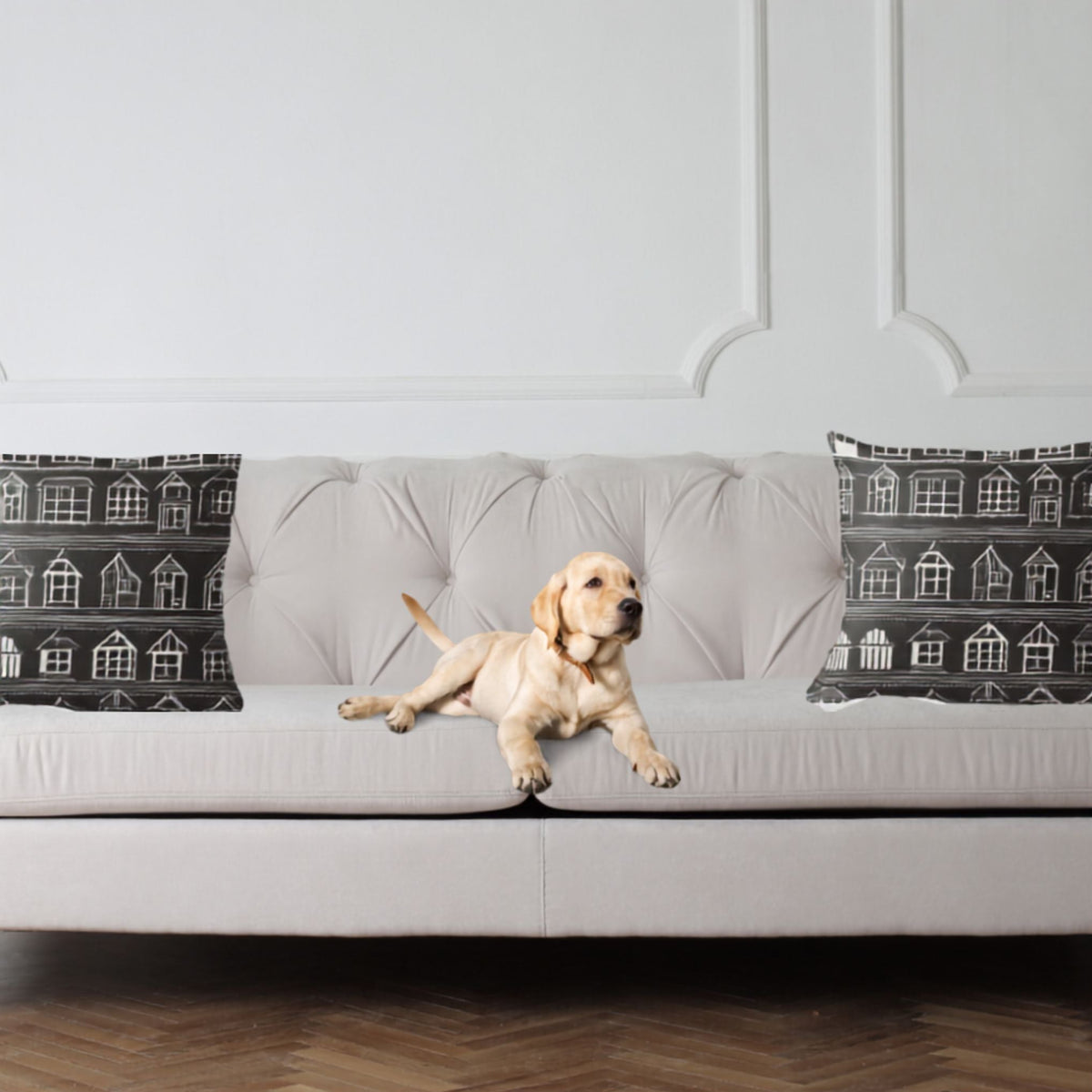 A light cream tufted sofa in a bright living room, with a yellow Labrador retriever lying on the seat cushion. Two black throw pillows with white house illustrations are placed on either side of the sofa.