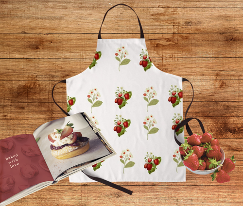 A Vintage inspired strawberry print apron on a wood background with a bowl of berries and a cookbook