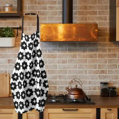 A black and white patterned kitchen apron with a star motif hangs on a wall in a rustic kitchen setting with a brick backsplash, copper stove hood, and wooden cabinets.