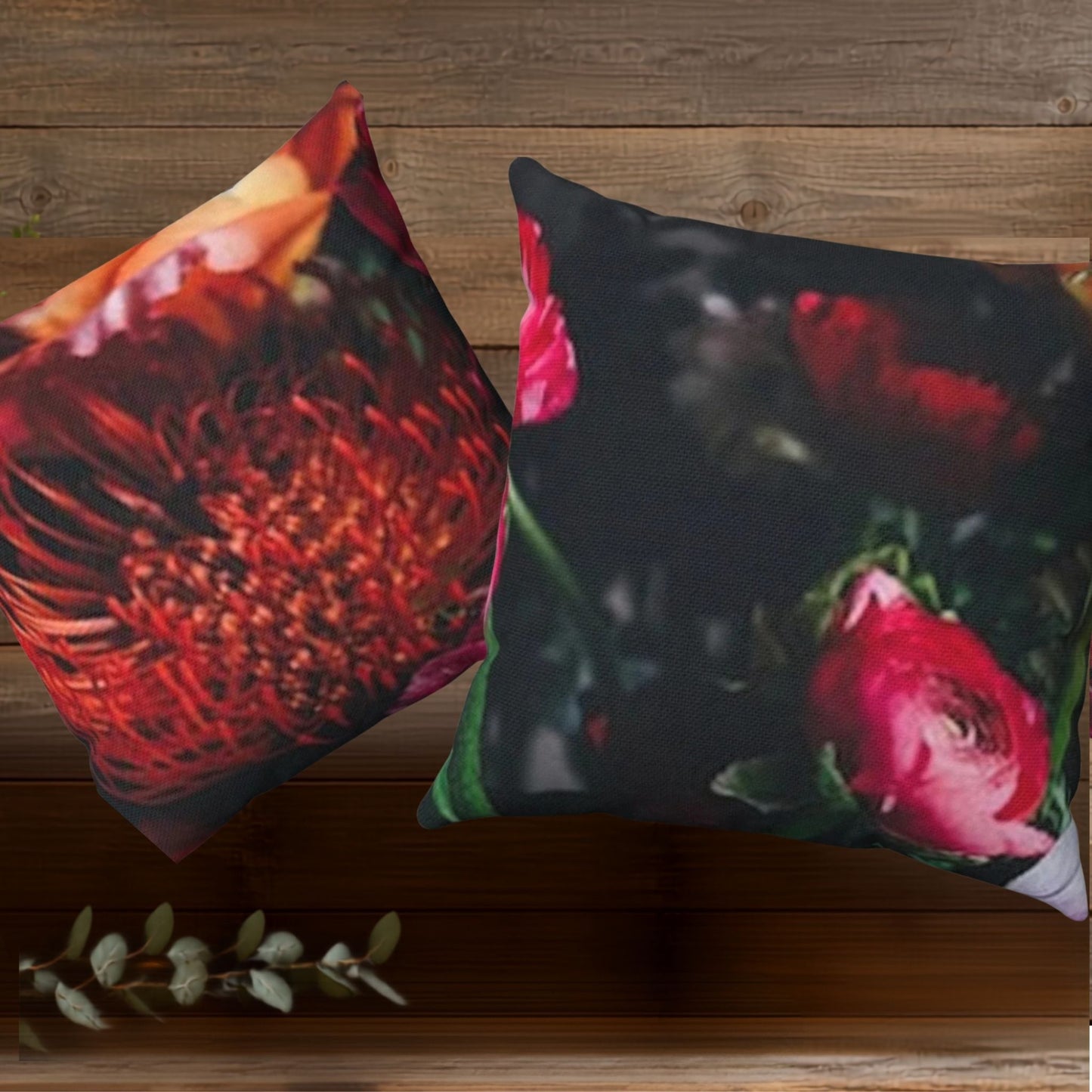 Two decorative throw pillows with floral prints displayed on a rustic wooden surface. One pillow features a large red protea flower on a dark background, and the other shows pink peony flowers on a deep, dark background.