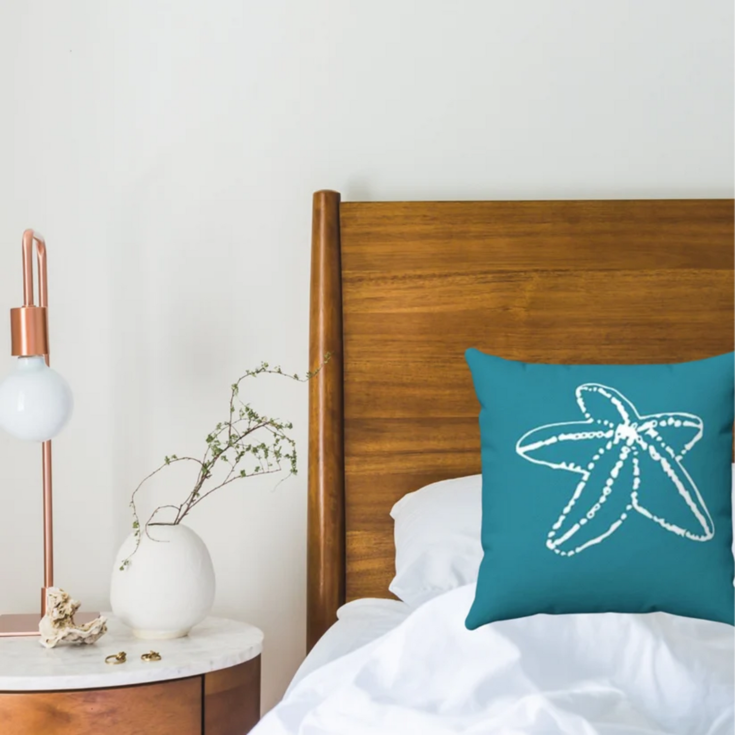 A teal blue throw pillow with a white starfish graphic printed on the front, styled on a bed with a wooden headboard and a bedside table with a lamp and vase.