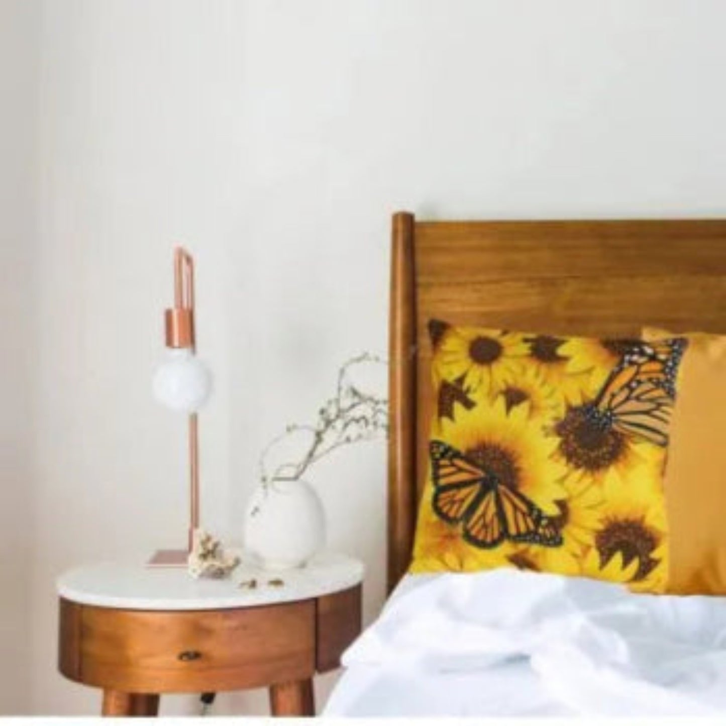 A close-up lifestyle shot of a bed with a wooden headboard, featuring a yellow sunflower and butterfly patterned pillow, white bedding, and a matching wooden nightstand with a copper and white table lamp.