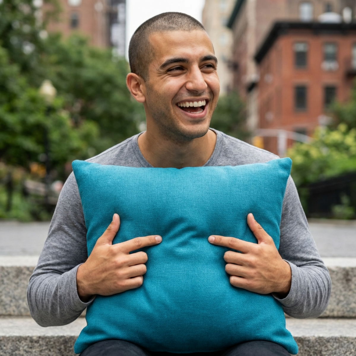 Man holding a teal pillow outdoors with buildings and greenery in the background