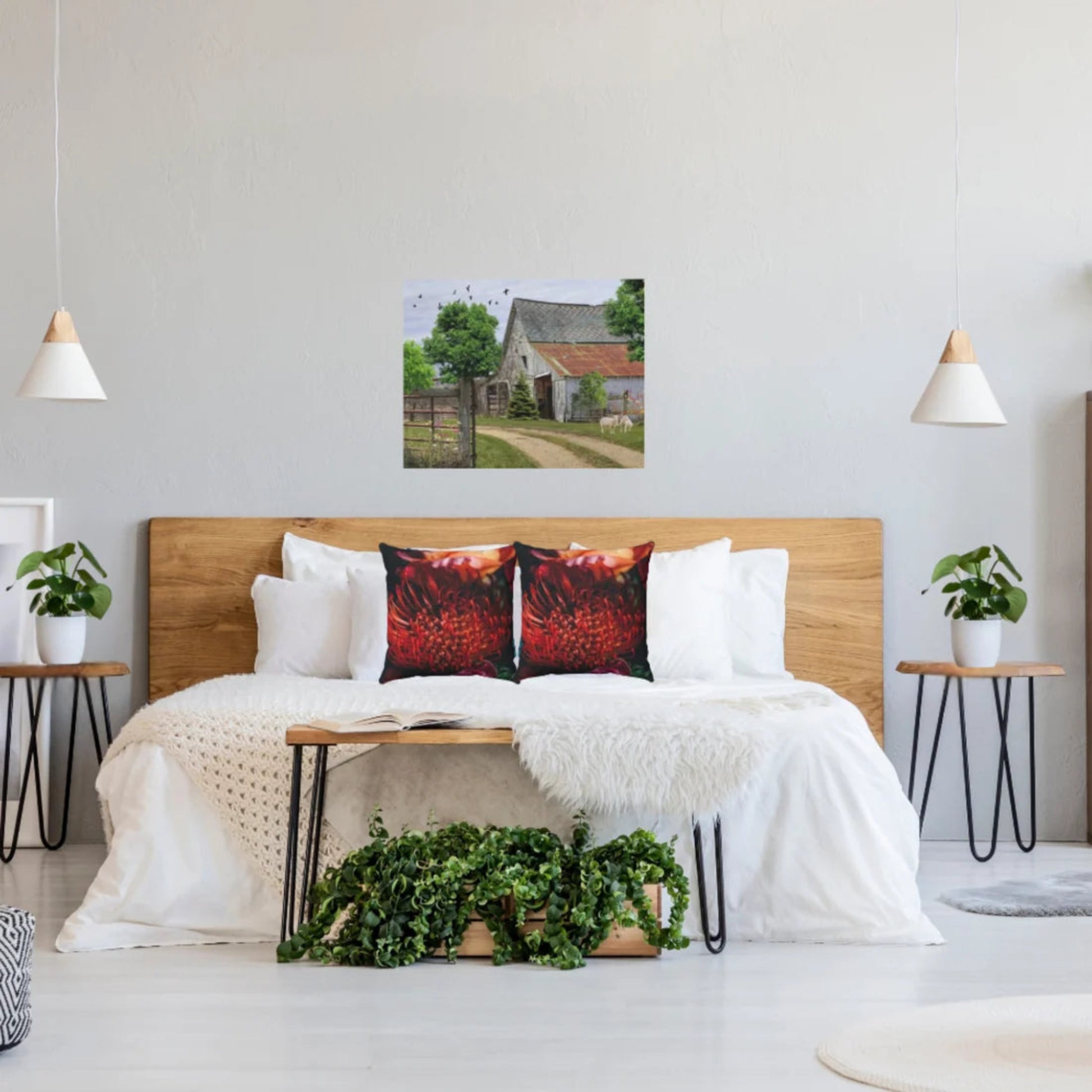 A bedroom scene with a bed dressed in white bedding and two deep orange-and-black floral pillows, beneath a wall art print showing a rustic barn and trees. The room has a light wood headboard, white walls, pendant lights, side tables, potted plants, and a bench with greenery.