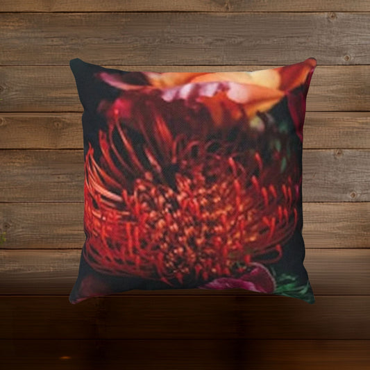 A square throw pillow featuring a close-up photographic print of a red protea flower, set against a rustic brown wooden plank background.
